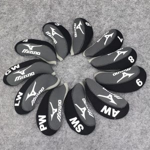 Puede incluir: Un conjunto de fundas para palos de golf negras y grises. Cada funda tiene un logotipo blanco de Mizuno y un número o abreviatura, incluyendo 4, 6, 7, 8, GW, LW, PW, MS y AW. Las fundas están dispuestas en un patrón circular.