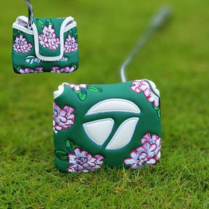 Puede incluir: Una funda de putter de golf verde con un diseño floral con flores rosas y blancas. La funda tiene un ribete blanco y el logotipo de TaylorMade en blanco. La funda está sobre una superficie de hierba verde.
