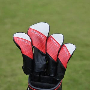 May include: A set of four golf club head covers. The covers are black, red, and white, with the word "Stealth" printed on each. The red portion has a honeycomb pattern. The covers are in a golf bag, with a green background.