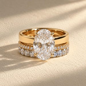Könnte beinhalten: Ein Goldring-Set bestehend aus zwei Ringen. Der obere Ring hat einen großen, oval geschliffenen, klaren Mittelstein, der von vier Krappen gehalten wird. Der untere Ring ist ein Pavé-Ewigkeitsring, besetzt mit vielen kleinen, runden Steinen.