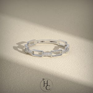 May include: A silver ring with a chain link design. The ring features rectangular links with small, sparkling stones set into some of the links. The ring is displayed on a neutral, textured surface.