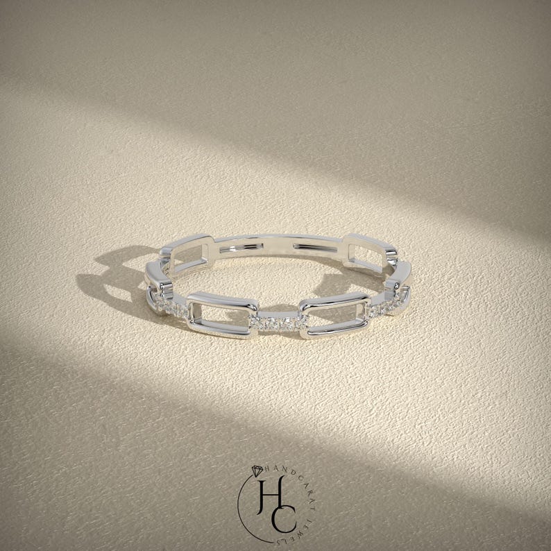 May include: A silver ring with a chain link design. The ring features rectangular links with small, sparkling stones set into some of the links. The ring is displayed on a neutral, textured surface.