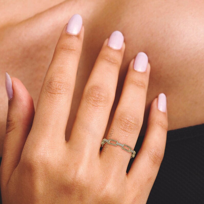 May include: A gold-colored chain link ring with small, clear stones set in the links. The ring is worn on a finger, with the hand and arm visible. The nails are painted a light pink color.