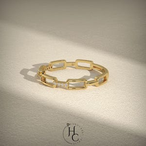 May include: A gold ring with a chain link design. The ring features rectangular links and small, sparkling stones set in a row. The ring is displayed on a neutral, textured surface, with a logo in the bottom right corner.