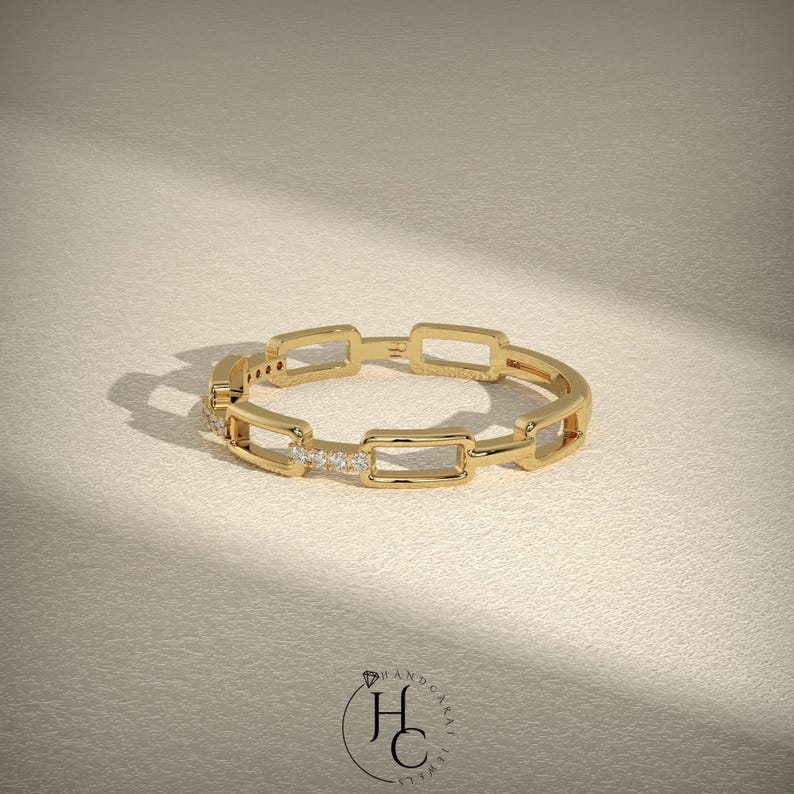May include: A gold ring with a chain link design. The ring features rectangular links and small, sparkling stones set in a row. The ring is displayed on a neutral, textured surface, with a logo in the bottom right corner.