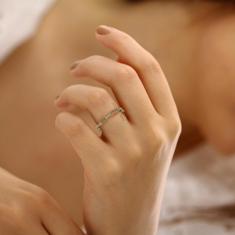 May include: A gold-colored chain link ring with small, clear gemstones. The ring is worn on a finger, showcasing its delicate design and sparkle. The ring's links are rectangular, creating a modern and elegant look.