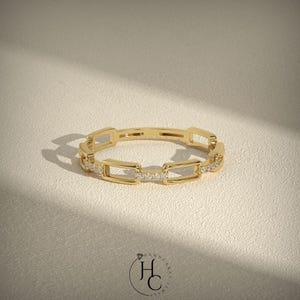 May include: A gold-colored ring with a chain link design. The ring features rectangular links with small, clear gemstones set in some of the links. The ring is displayed on a neutral-colored surface, with soft shadows.
