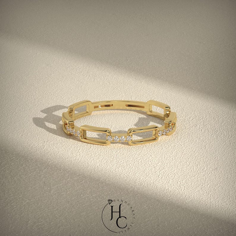 May include: A gold-colored ring with a chain link design. The ring features rectangular links with small, clear gemstones set in some of the links. The ring is displayed on a neutral-colored surface, with soft shadows.