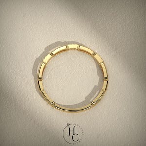 May include: A top-down view of a gold ring with a rectangular design. The ring is made of gold and has a series of rectangular segments around the band. The ring is set against a neutral background.