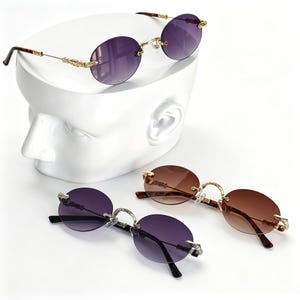 May include: Oval sunglasses with tinted purple and brown lenses. The frames have gold and silver detailing. One pair rests on a white head sculpture, with two more below.