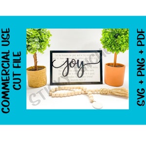 May include: A black framed sign with the word "joy" in a decorative font. The sign has a white background with a bible verse printed on it. The sign is sitting on a white surface with a wooden bead garland and a small potted plant.
