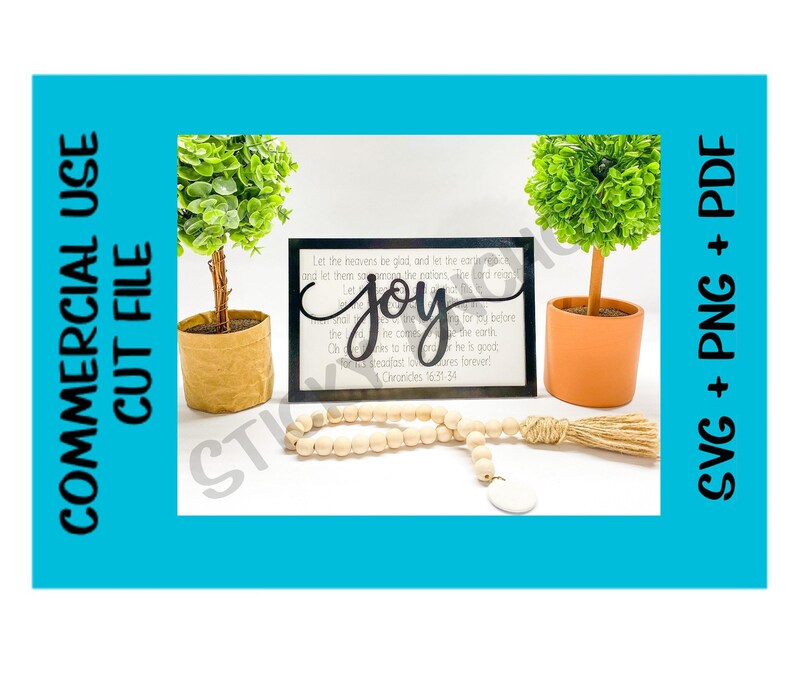 May include: A black framed sign with the word "joy" in a decorative font. The sign has a white background with a bible verse printed on it. The sign is sitting on a white surface with a wooden bead garland and a small potted plant.