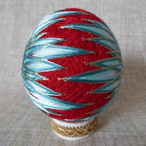 May include: An egg-shaped ornament with a red base, decorated with teal and gold thread in a leaf pattern. The ornament is on a white base with gold trim, demonstrating detailed craftsmanship. The ornament is approximately 8 cm tall.