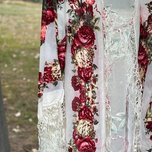 May include: A sheer white floral kimono with a vibrant red rose pattern and cream-colored lace trim. The long sleeves and open front design create a flowing silhouette. The kimono is a lightweight, bohemian style garment.