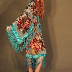 May include: A light blue floral kimono with a turquoise trim and fringe. The kimono features a large floral pattern in shades of red, burgundy, and yellow. The model is wearing black high heels.