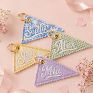 Peut inclure: Quatre porte-clés en tissu triangulaires avec des anneaux en métal doré. Chaque pièce présente des bords festonnés et des noms brodés en blanc : Sophia en bleu floral, Alex en vichy vert, Jack en rayures jaunes et Mia en tissu violet clair.