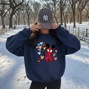 May include: Navy blue sweatshirt featuring a graphic of Spider-Man in a Mickey Mouse-inspired design, holding balloons with various Disney characters. The sweatshirt also has images of Mickey Mouse-themed treats. A gray baseball cap with a white "NY" logo is also visible.