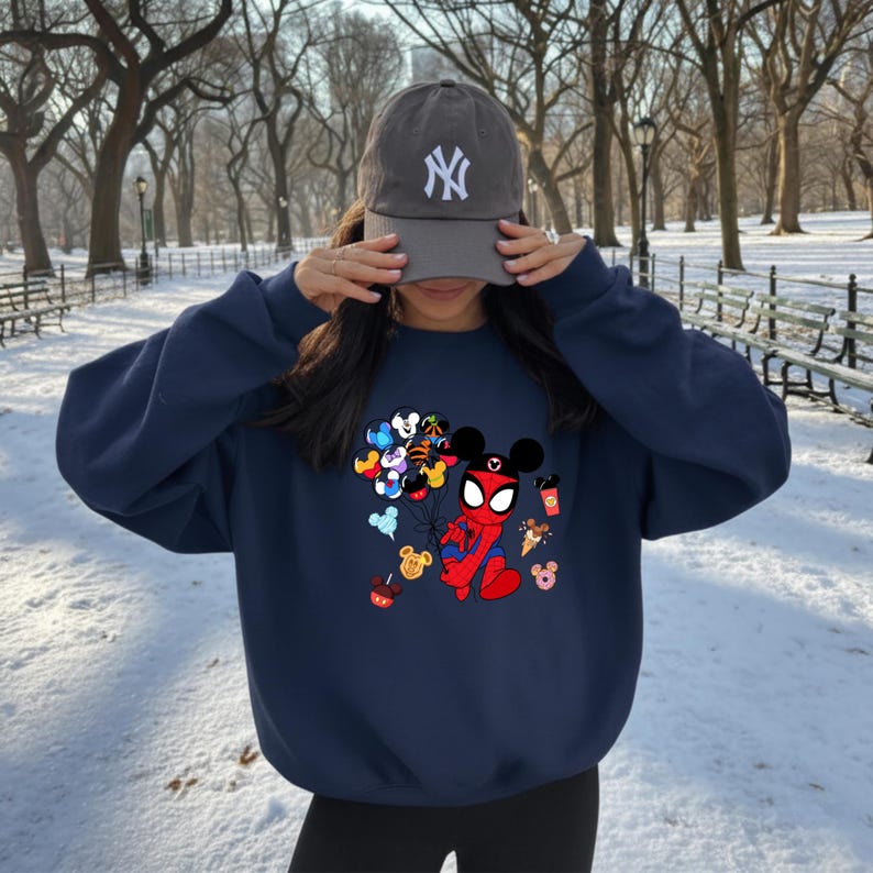 May include: Navy blue sweatshirt featuring a graphic of Spider-Man in a Mickey Mouse-inspired design, holding balloons with various Disney characters. The sweatshirt also has images of Mickey Mouse-themed treats. A gray baseball cap with a white "NY" logo is also visible.