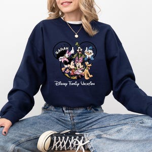 May include: Navy blue sweatshirt featuring a Disney family graphic. The graphic includes Mickey and Minnie Mouse, Goofy, Donald Duck, and other characters. The word "SARAH" is above the characters, and "Disney Family Vacation" is below.