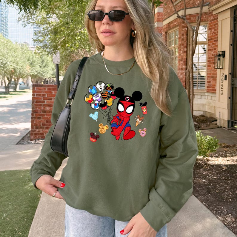 May include: Olive green sweatshirt featuring a cartoon illustration of a Spider-Man character with Mickey Mouse ears holding balloons. The graphic includes various Disney-themed icons. The sweatshirt is long-sleeved.