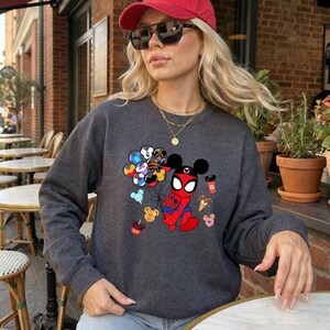 May include: Dark gray sweatshirt featuring a cartoon mashup of Spider-Man and Mickey Mouse. The design includes Mickey Mouse ears, Spider-Man's suit, and balloons with Disney character designs. Additional Disney-themed icons are scattered around the main graphic.