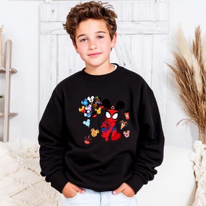 May include: A black sweatshirt featuring a colorful graphic of Mickey Mouse balloons, Spider-Man, and various Disney-themed treats. The design includes red, blue, yellow, and black elements. The sweatshirt is worn by a young person.