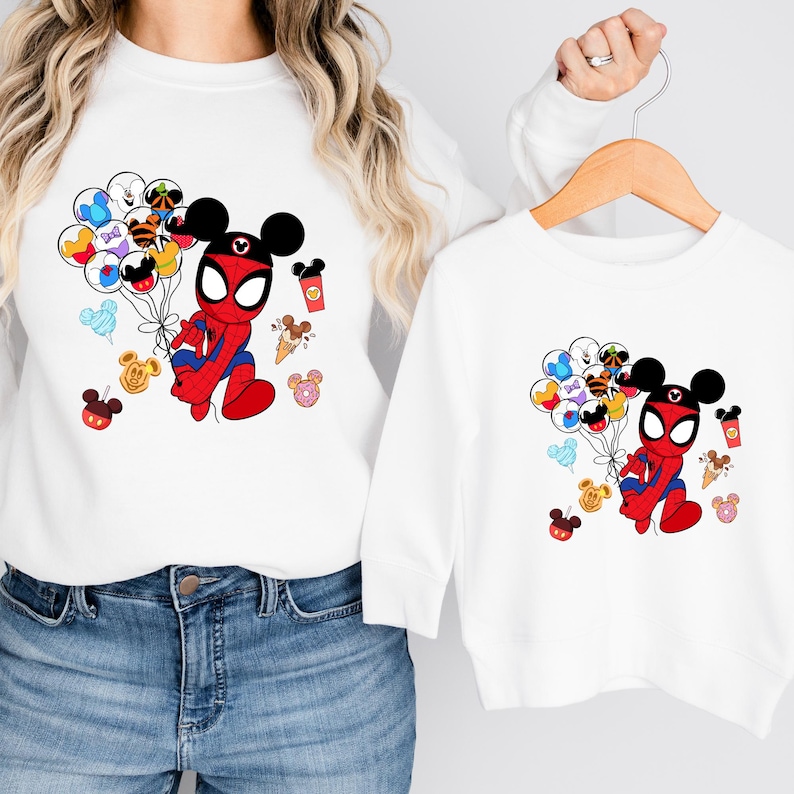 May include: White sweatshirts featuring a cartoon Spider-Man character with Mickey Mouse ears, holding balloons with Disney character faces. Additional Disney-themed graphics include Mickey Mouse heads, ice cream cones, and coffee cups. The sweatshirts are displayed on a hanger.