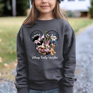 May include: Dark gray sweatshirt featuring a Disney family graphic. The graphic includes Mickey and Minnie Mouse, Donald Duck, Goofy, and Pluto. The name "EMMA" is above the characters, and the text "Disney Family Vacation" is below.