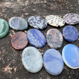Puede incluir: Un surtido de piedras preciosas ovaladas pulidas en varios colores, incluyendo verde, azul, rojo y gris. Las piedras tienen patrones y texturas únicos, exhibidas sobre una superficie texturizada de color marrón oscuro.