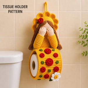 May include: A yellow crocheted tissue holder pattern featuring a figure with brown braided hair, a yellow bonnet, and hands covering the face. The holder has a cylindrical base decorated with red and green floral motifs and a white daisy.