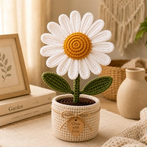 May include: A crocheted daisy flower with white petals and a yellow center, featuring two green leaves on a stem. The flower is planted in a beige crocheted pot with a brown top, decorated with a twine bow and a wooden tag reading Grow with Love.