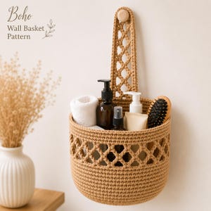 May include: A tan crocheted wall basket with a diamond-patterned strap hanging from a wooden peg. The basket contains a white rolled towel, a dark brown pump bottle, a small spray bottle, a white tube, and a black hairbrush. Text reads: Boho Wall Basket Pattern.