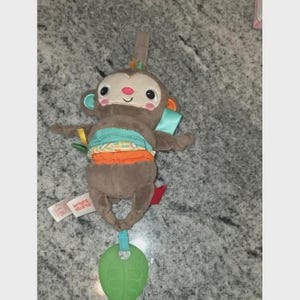 Op de afbeelding: Een bruin, pluche aap-speeltje met een lachend gezicht, blauwe en oranje accenten en een groene, bladvormige bijtring. Het speelgoed heeft een lus om op te hangen en een label met "Bright Starts".
