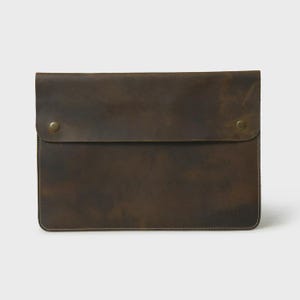 May include: A brown leather laptop sleeve with a flap closure secured by two brass-colored snap buttons. The sleeve has a distressed finish and visible stitching along the edges, suggesting a handcrafted design. The overall aesthetic is rustic and durable.