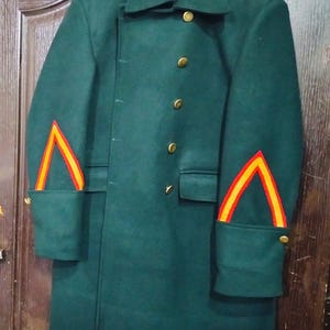 May include: A dark green military-style coat with gold buttons and red and yellow chevron accents on the sleeves. The coat has a classic collar and epaulettes. The coat is hanging on a hanger.