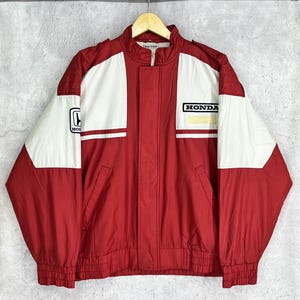 May include: A red and white Honda racing jacket with a zip-up front and a stand-up collar. The jacket features the Honda logo on the chest and sleeve. The jacket has a ribbed hem and cuffs.