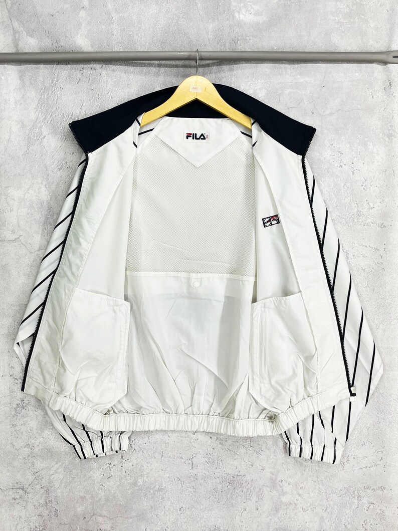 May include: White and black Fila jacket with a black collar and black vertical stripes on the sleeves. The jacket has a mesh lining and two front pockets. The Fila logo is visible on the chest and sleeve.