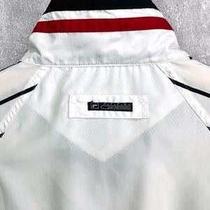 May include: White athletic jacket with black and red accents. The collar features black, red, and white stripes. The jacket has a small rectangular tag that reads "performance".