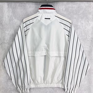 May include: White jacket with black vertical stripes on the sleeves. The jacket has a stand-up collar with red and navy blue accents. The back of the jacket is plain white with a small logo near the collar. The jacket has a drawstring hem.