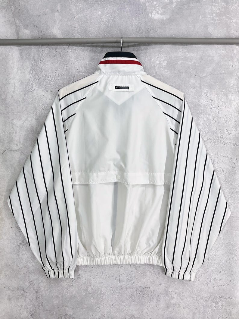 May include: White jacket with black vertical stripes on the sleeves. The jacket has a stand-up collar with red and navy blue accents. The back of the jacket is plain white with a small logo near the collar. The jacket has a drawstring hem.