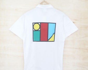 May include: White polo shirt with a colorful geometric design on the back. The design features a yellow circle, red rectangle, blue triangle, and green square, all outlined in black. The shirt is hanging on a wooden hanger.