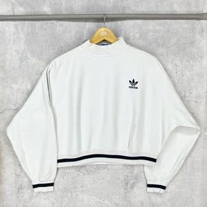 May include: White cropped sweatshirt with a high neck and long sleeves. Features the Adidas logo on the chest. The bottom hem and cuffs have a black and white striped design. The sweatshirt is hanging on a wooden hanger.