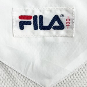 May include: Close-up of a white fabric with a stitched label. The label features the brand name "FILA" in navy blue with a red accent, and the number "-100" in red. The fabric has a mesh-like texture.
