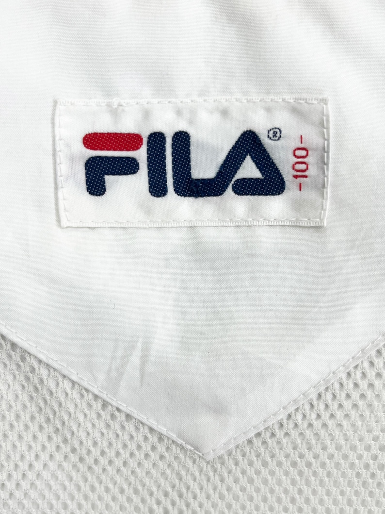 May include: Close-up of a white fabric with a stitched label. The label features the brand name "FILA" in navy blue with a red accent, and the number "-100" in red. The fabric has a mesh-like texture.