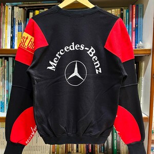 May include: Black sweatshirt with red and black color block design. The back features the Mercedes-Benz logo in white. The sleeves have red accents and text that reads "st bl PARK" and "mpion".