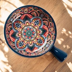 以下が含まれることがあります： 青いセラミック製の丸いボウルで、持ち手が付いており、詳細でカラフルな花のマンダラ模様が特徴です。ボウルの内側には、赤、黄、青、緑の色合いの鮮やかなパターンが施されています。ボウルは木製の表面に置かれています。