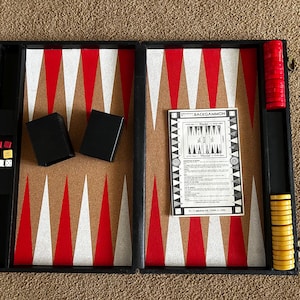 May include: An open black backgammon board game set featuring a tan playing surface with red and white triangular points. The set includes two black dice cups, a white instruction booklet titled BACKGAMMON, red and yellow checkers, and dice.