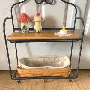 May include: A black metal two-tiered shelf with a wooden top shelf and a woven basket on the bottom shelf. The shelf has hooks on the bottom and is decorated with two ceramic mushroom figurines and a small candle.