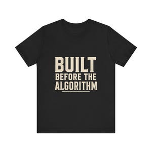 May include: A black short-sleeved t-shirt featuring the text BUILT BEFORE THE ALGORITHM printed in a bold, off-white, distressed serif font across the centre of the chest, with a horizontal line positioned directly underneath the text.