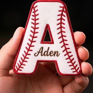 May include: White baseball-themed patch in the shape of the letter "A", with red stitching and trim. The name "Aden" is embroidered in brown. The patch is held against a dark background.